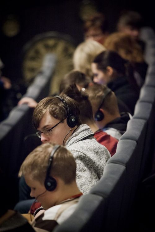 Nuotraukoje - žiūrovai sėdi su ausinėm ir laukia spektaklio su garsiniu vaizdavimu Nuotraukoje - žiūrovai sėdi su ausinėm ir laukia spektaklio su garsiniu vaizdavimu