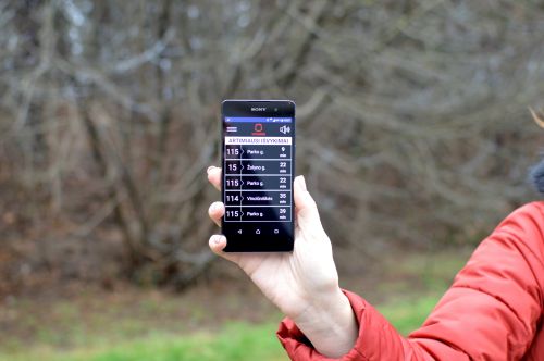 Nuotraukoje - ranka, laikanti išmanųjį telefoną su ekrane atversta TRANSPORTO BALSO programėle, rodančia artimiausius išvykimus Nuotraukoje - ranka, laikanti išmanųjį telefoną su ekrane atversta TRANSPORTO BALSO programėle, rodančia artimiausius išvykimus
