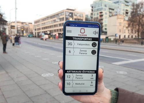 Nuotruakoje - mobilusis telefonas, su įjungta TRANSPORTO BALSO programėle, miesto fone