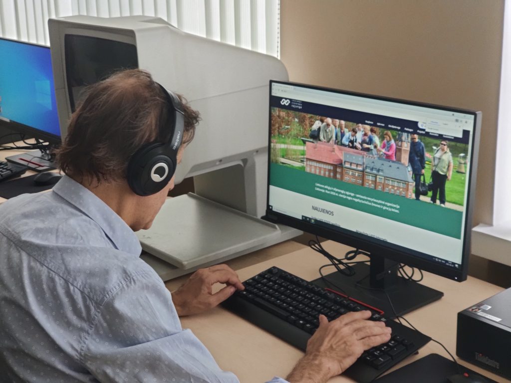 Nuotraukoje - vyras sėdi prie kompiuterio ekrano su ausinėmis, rankos uždėtos ant klaviatūros. Monitoriaus ekrane matosi svetainės www.lass.lt pirmasis puslapis.