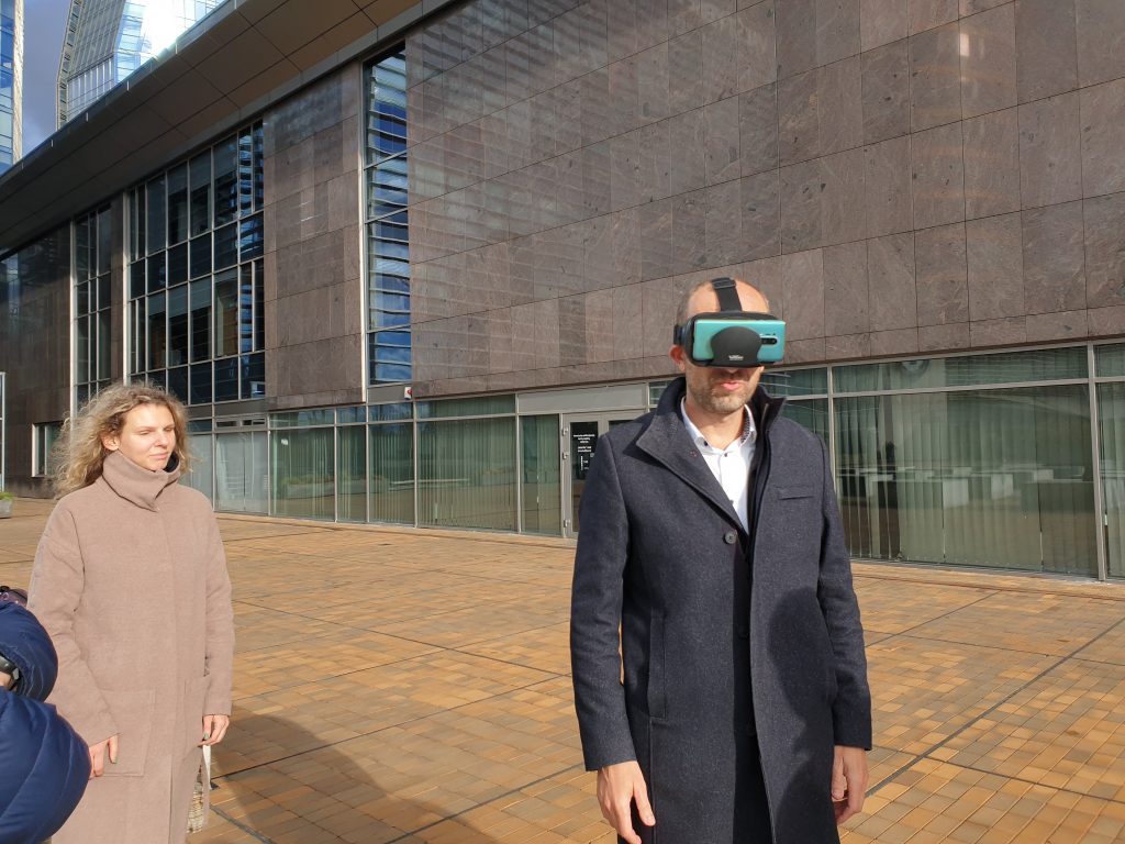 Nuotraukoje - vyras ir moteris, kurie stovi prie pilko pastato. Vyras užsidėjęs ant galvos virtualios realybes akinius, kurie simuliuoja įvairius regos sutrikimus. 