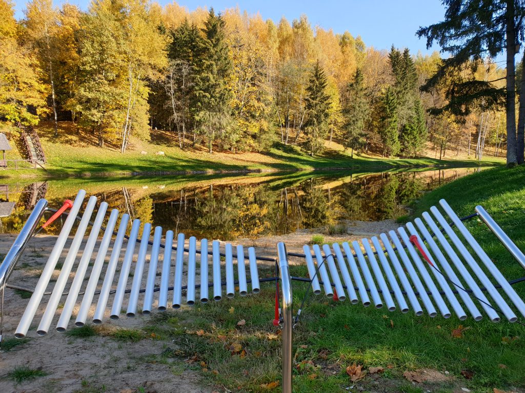 Nuotraukoje - iš įvairaus ilgio vamzdelių sudarytas instrumentas, kuris yra rudeninių medžių ir vandens telkino fone. 
