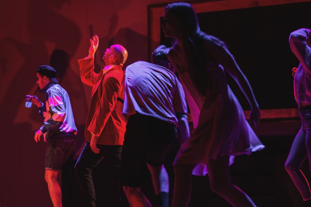 Nuotraukoje - blankiai apšviestoje scenoje juda 5 žmonės. jie apšviesti raudonais ir violetiniais žibintais, siluetai neryškųs. 