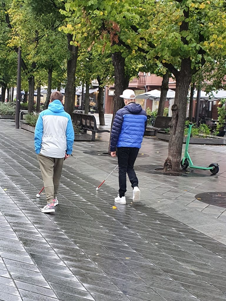 Nuotraukoje - vyras ir moteris eina pėsčiųjų taku, naudodamiesi baltosiomis lazdelėmis. Jiems iš dešinės auga medžiai, matosi suoliukai ir kavinių skėčiai.