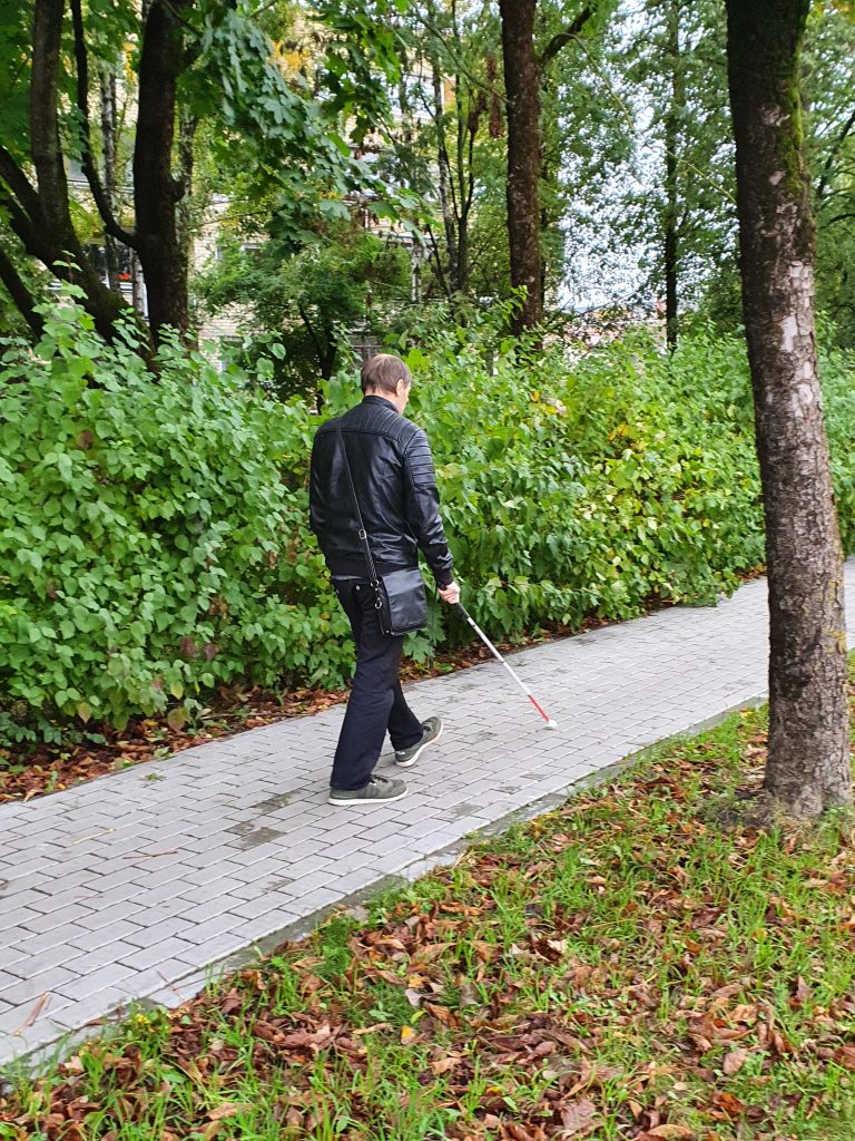 Nuotraukoje - vyresnio amžiaus vyras eina šaligatviu miškingoje vietovėje, naudodamasis baltąja lazdele. 
