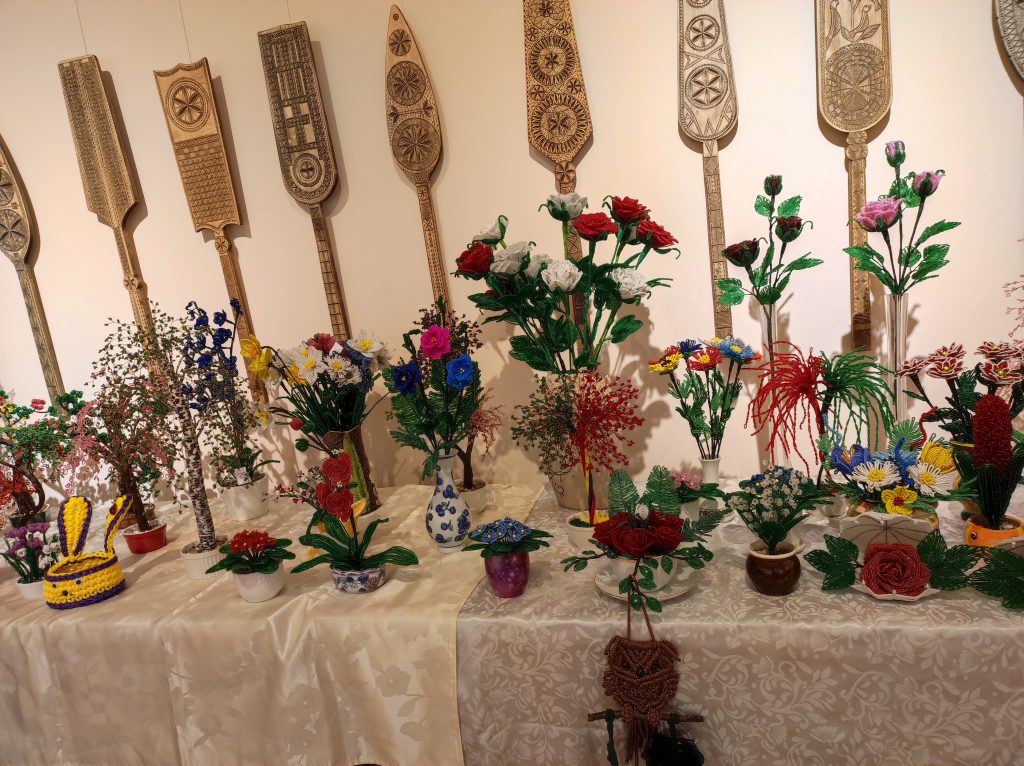 Nuotraukoje - išdėliotos ant stalo dirbtinių gėlių puokštės, ant sienos kabo įvairaus dizaino medinės verpstės.